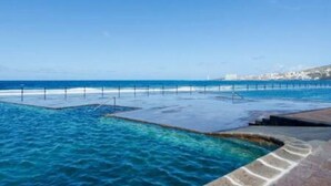 A coastal swimming pool with clear blue water, a stone deck, and a view of the sea and distant buildings. - Apartamento Piscinas Naturales (San Cristóbal de La Laguna)