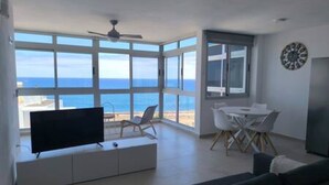 A modern living room with a large TV, a dining area, and a view of the ocean. - Apartamento Piscinas Naturales (San Cristóbal de La Laguna)
