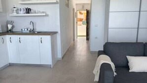A modern kitchen with white cabinets, a countertop, and a sink. There is a dark-colored sofa with a white throw blanket in the living area. - Apartamento Piscinas Naturales (San Cristóbal de La Laguna)