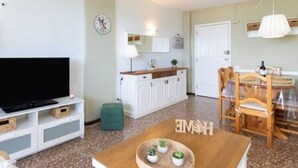 A modern living room with a flat-screen TV, a wooden coffee table with decorative items, a dining area with a wooden table and chairs, and a kitchen area with white cabinets and a countertop. - Coral Sunsent View With 1 Bedrooms and 1 5 Bathrooms (Tacoronte)