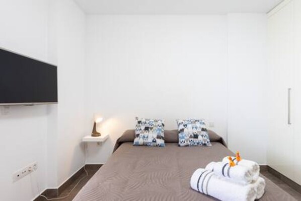 Une chambre d’hôtel avec un lit, une table de chevet, un téléviseur fixé au mur et une petite étagère avec une lampe.