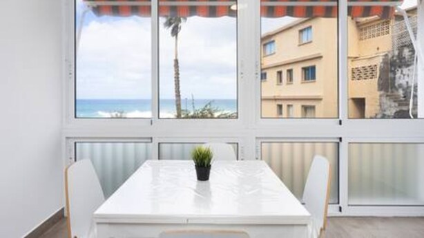A room with a white table and chairs, a potted plant, and a view of the sea and buildings through large windows. - Apartamento Brisa del Mar (Tacoronte)