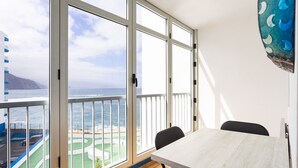 Una habitación con un gran ventanal que ofrece vistas al mar y una montaña distante. Hay una mesa con sillas y un reloj en la pared.
