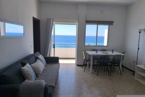 A room with a sofa, a dining table with chairs, and a view of the sea through a window. - La Caleta Reviva Canarias (San Cristóbal de La Laguna)