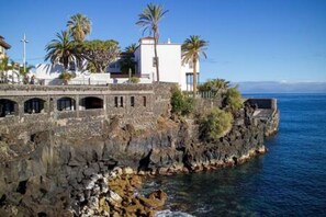 A coastal building with a stone facade, palm trees, and a view of the sea. - Apartamento Atardecer Martianez (Puerto de la Cruz)