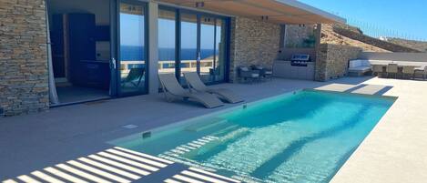 Un espace piscine extérieur moderne, doté de chaises longues, d’un mur en pierre et offrant une vue sur l’océan.
