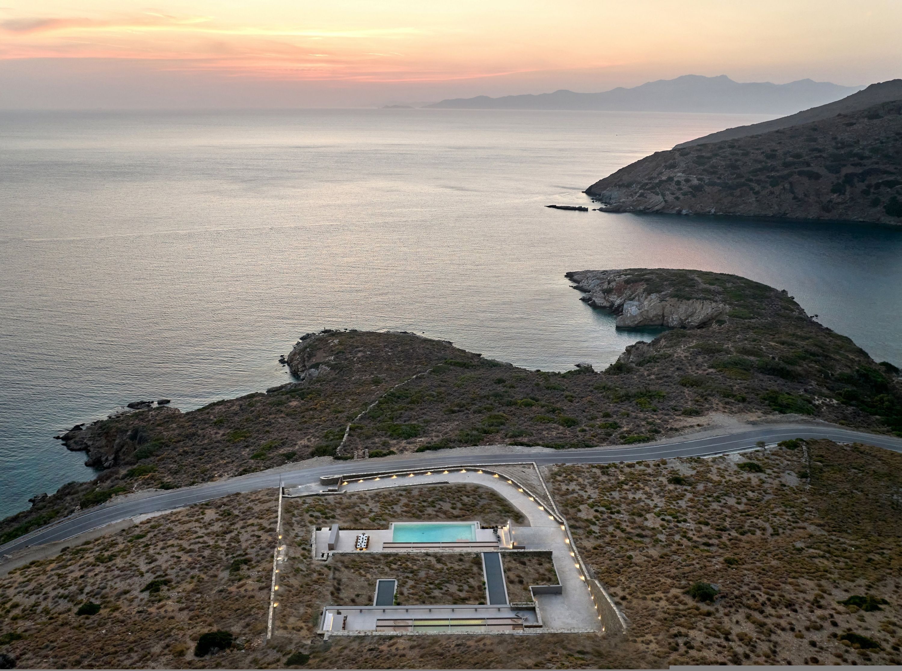 An aerial view of a coastal property with a swimming pool, a road, and rocky terrain.