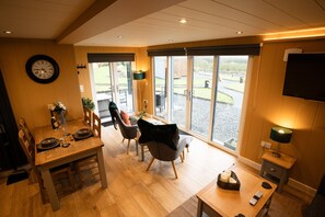 A dining area with a wooden table set for four, a clock on the wall, and a television on the wall. - Luxury Cabin with Hot Tub & Hill Views (Newton Stewart)