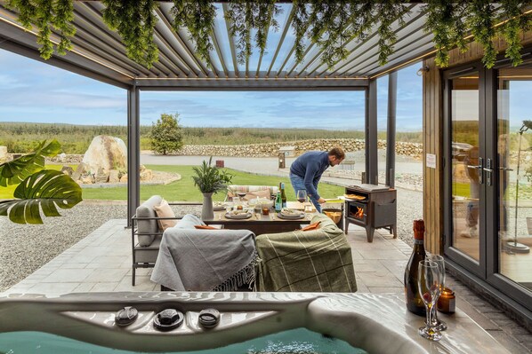 A covered outdoor seating area with a hot tub, a table set for dining, and a person tending to a wood-fired oven.