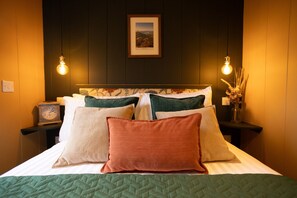 A neatly made bed with a green quilt, multiple pillows in various colors, a framed picture on the wall, and two wall-mounted light fixtures.