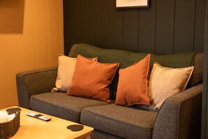 Un sofá gris con tres almohadas, un control remoto y una vela sobre una mesita de madera.