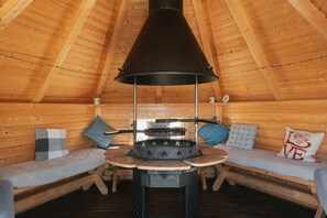 Una cabaña acogedora de madera con un fogón central, dos bancos de madera con cojines y un techo de madera inclinado.