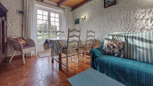 Una habitación rústica con techo de madera, pared de piedra, suelo de madera, un sofá azul, una mesa de comedor con sillas y una ventana con cortinas.