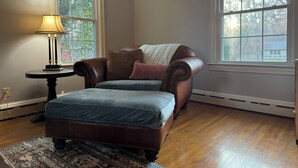 A room with a leather armchair, a matching ottoman, a lamp, and two windows with views of trees.