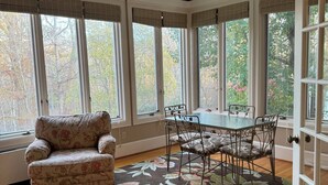 A room with a floral-patterned rug, a floral-patterned sofa, a dining table with chairs, and large windows with blinds.