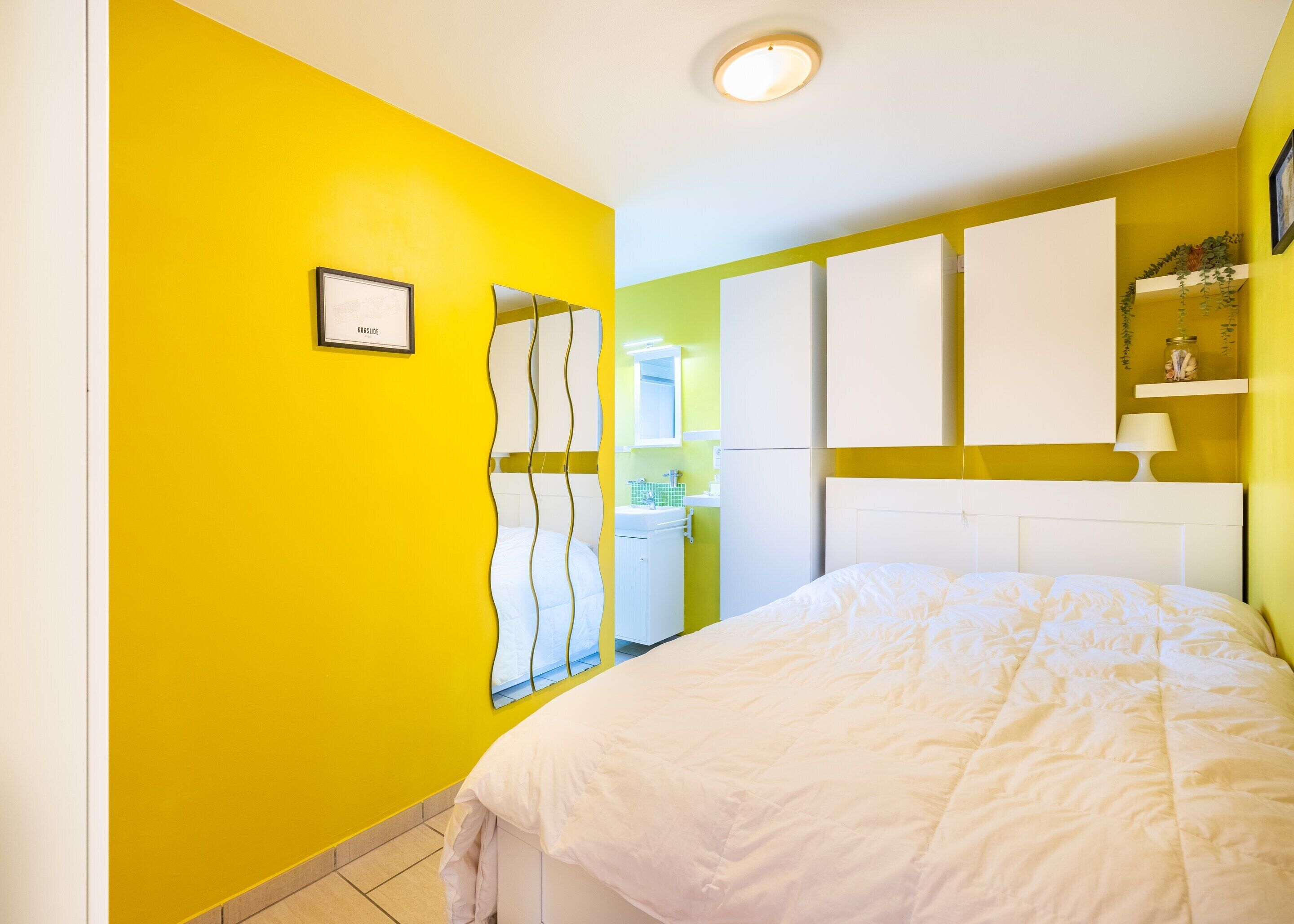 Quarto com parede amarela, cama branca e armário branco com espelho.