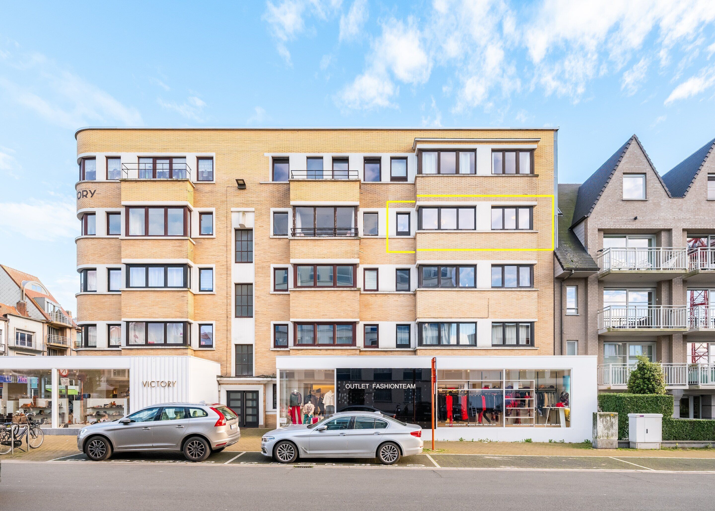 Un immeuble à plusieurs étages doté d’une architecture moderne, caractérisé par un mélange de façades en brique et en verre, et un espace commercial au niveau du rez-de-chaussée avec des devantures de magasins bien visibles.