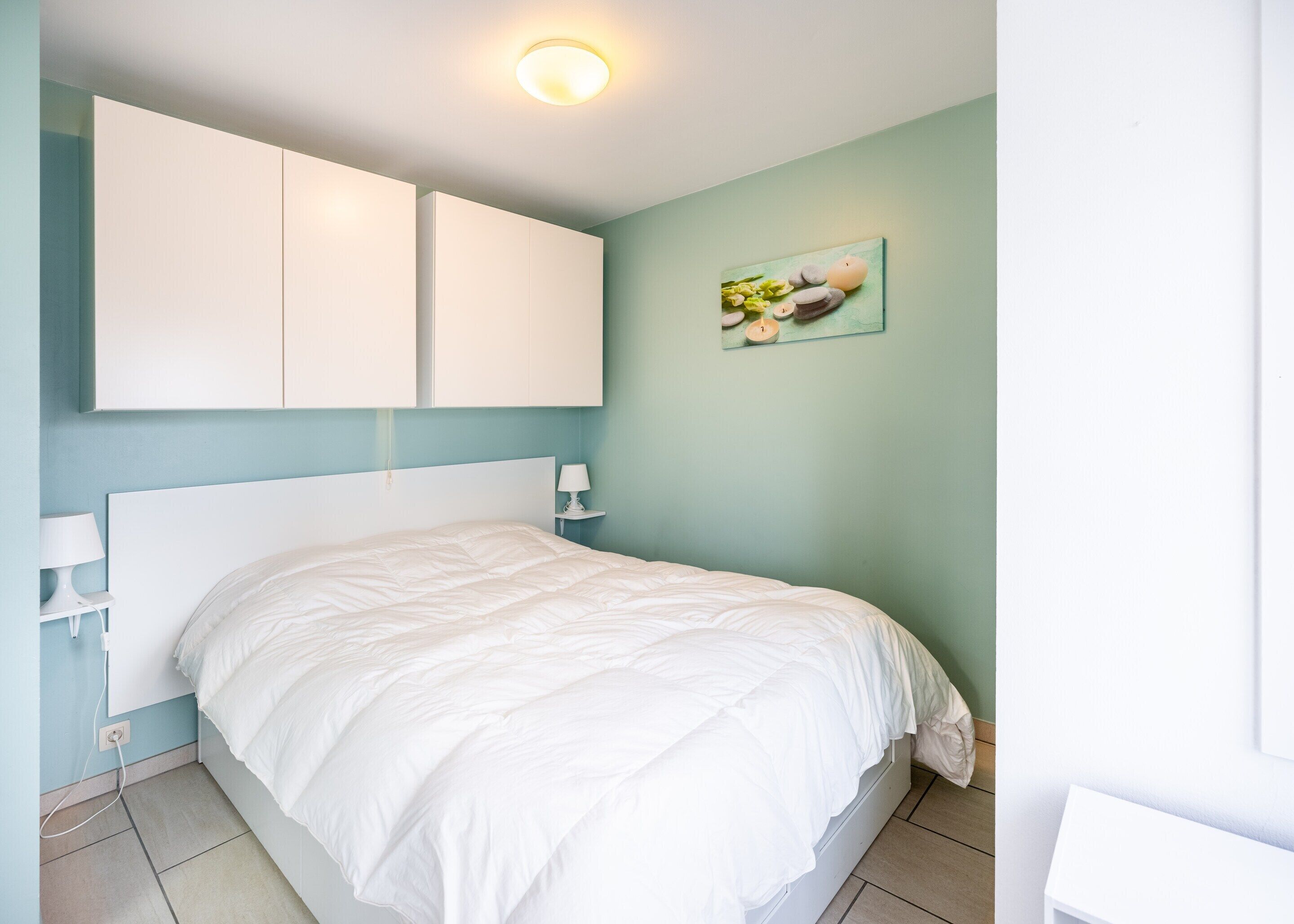 A bedroom with a white bed, two bedside tables with lamps, a ceiling light, and a wall-mounted artwork.