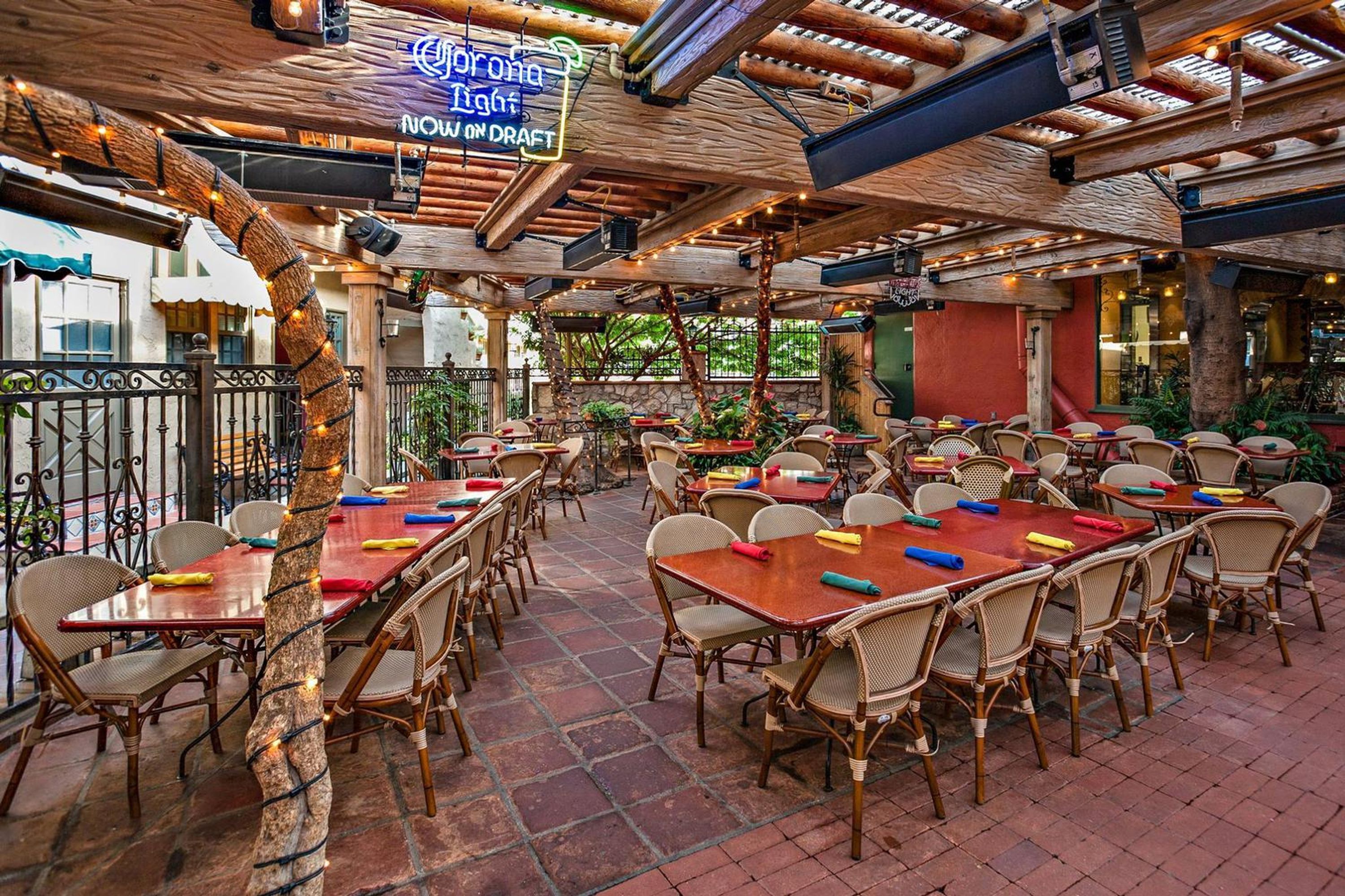 An outdoor dining area with wooden tables and wicker chairs, surrounded by a rustic wooden ceiling and brick flooring.