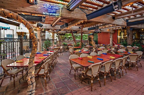 An outdoor dining area with wooden tables and wicker chairs, surrounded by a rustic wooden ceiling and brick flooring.