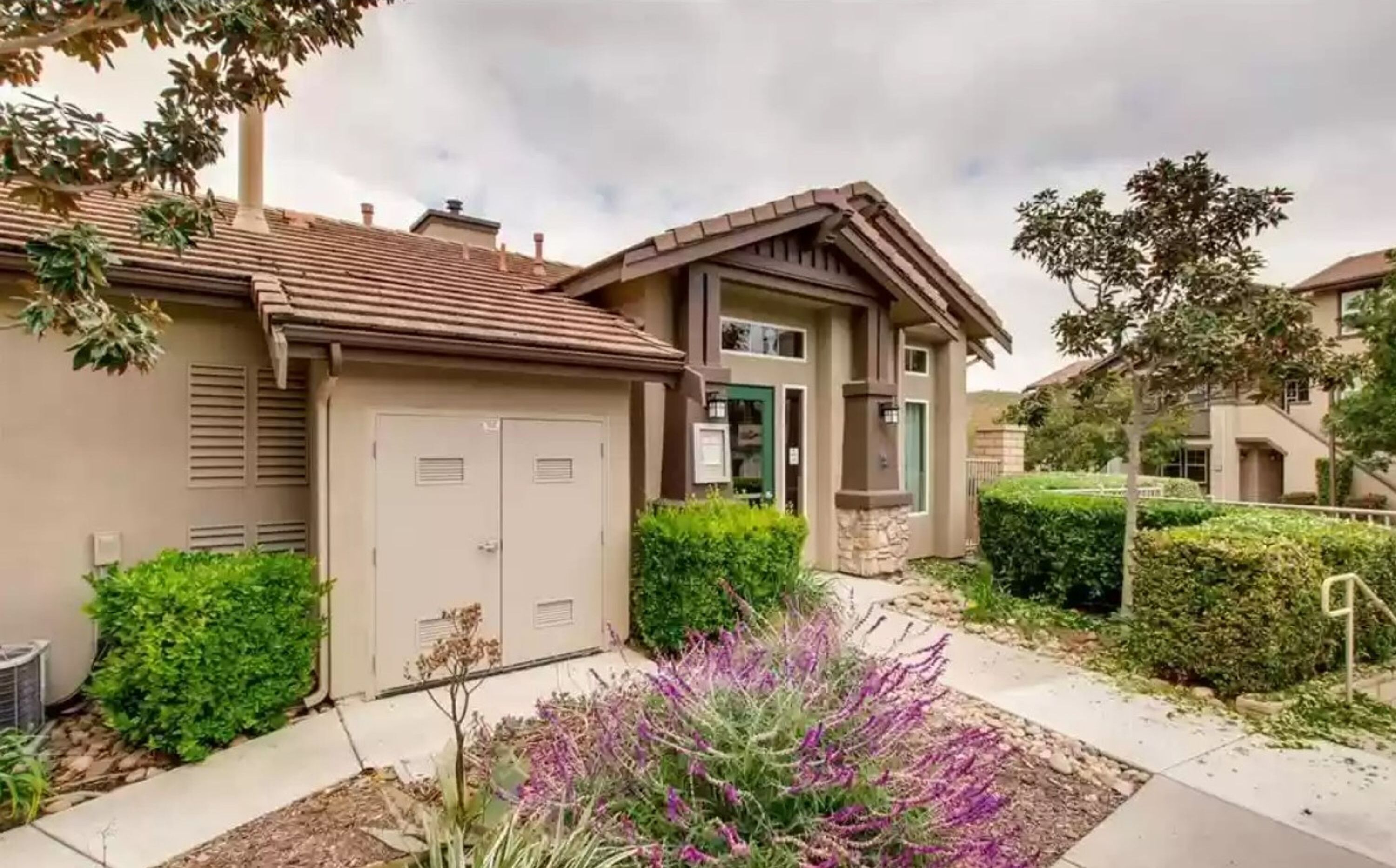 A residential building with a two-car garage, a front door, and a small front yard with purple flowers.