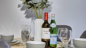 A dining table set with white bowls, plates, and wine glasses, accompanied by two bottles of wine and a vase with green foliage. - Lovely Flat in Kiln Close With Free Parking (Gloucester)