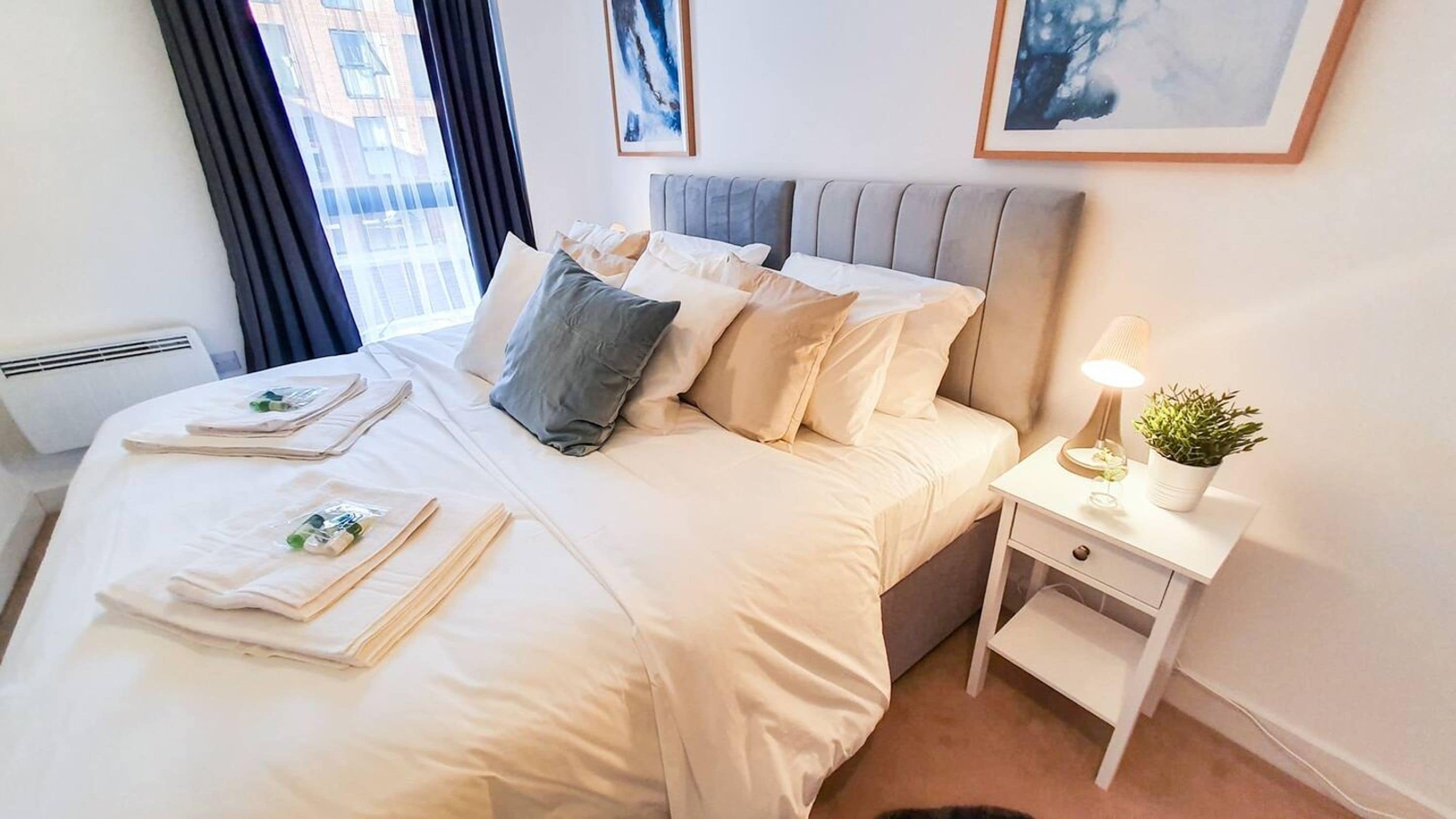 Een opgemaakt bed met kussens en een nachtkastje met een lamp en een plant.