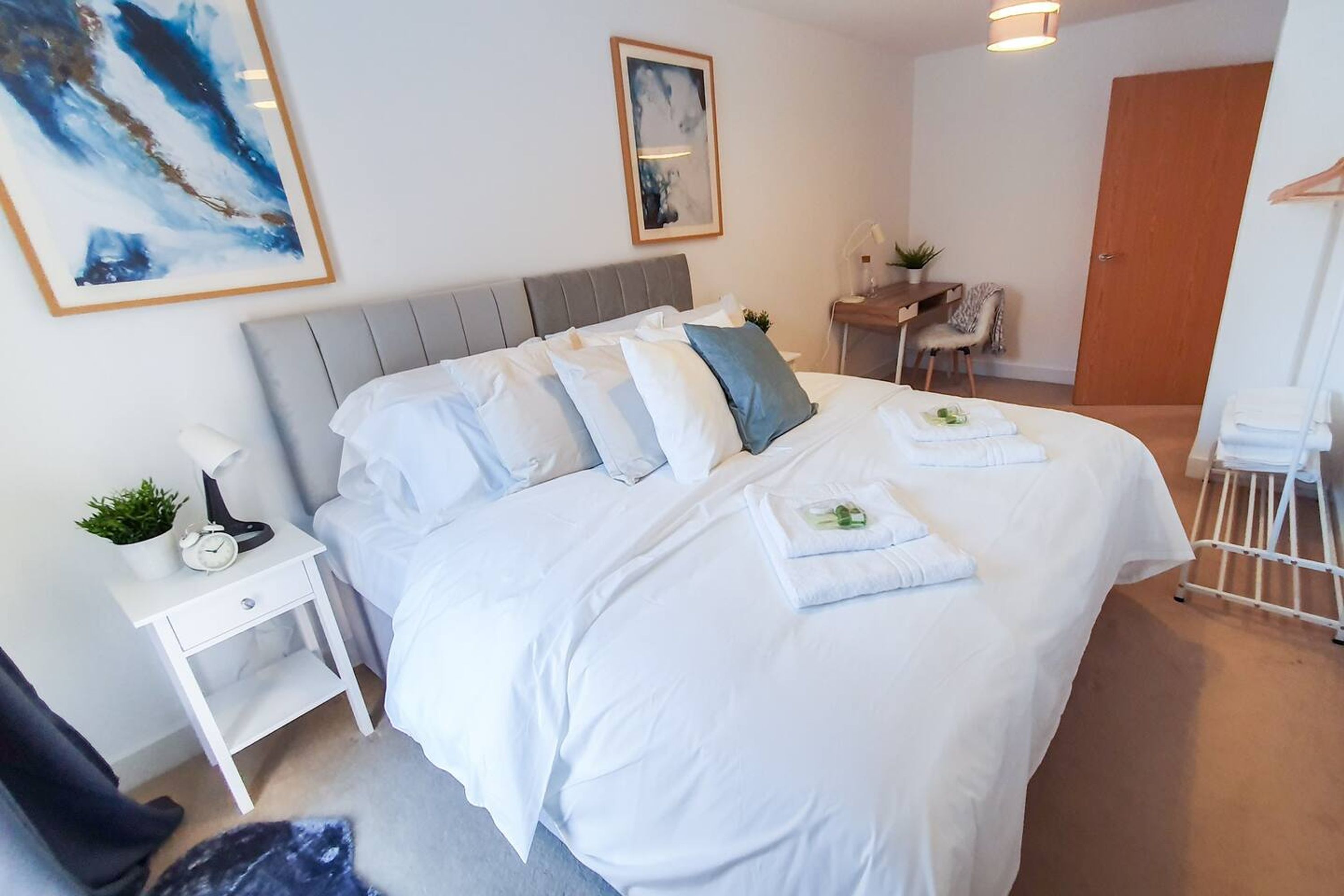 Een net opgemaakt bed met witte lakens, een nachtkastje met een lamp en een klok, en een klein plantje op de nachtkast.