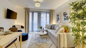 A modern living room with a sofa, a coffee table, a TV mounted on the wall, and a large window with curtains. - Lovely Flat in Kiln Close With Free Parking (Gloucester)