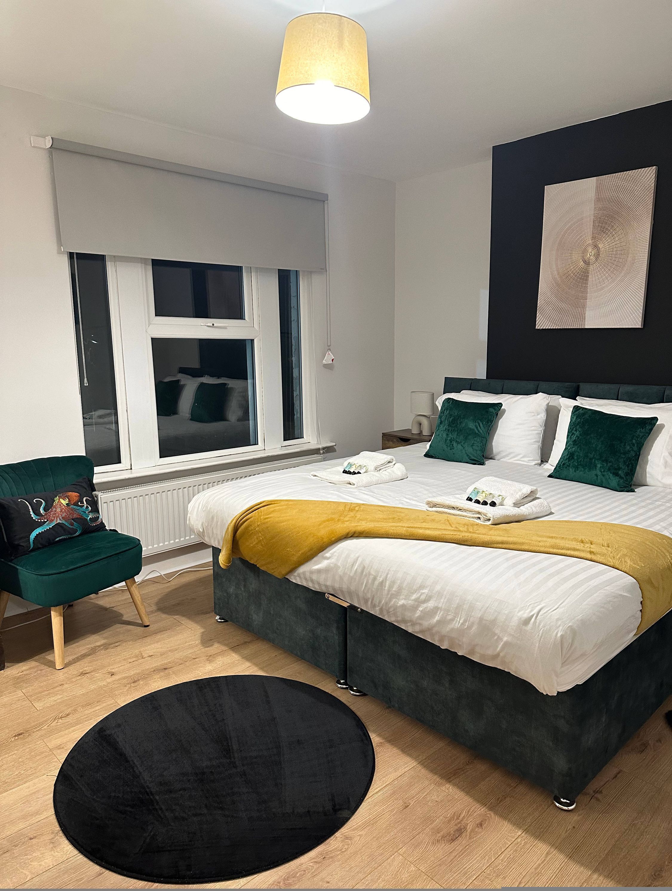 Un dormitorio moderno con una cama grande, un sillón verde, un tapete redondo negro y una ventana con persianas.