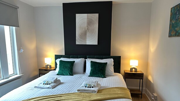 A neatly made bed with white and green pillows, a gold throw blanket, and a white towel on the side. There are two bedside tables with lamps, a black headboard, and a painting on the wall.