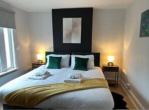 A neatly made bed with white and green pillows, a gold throw blanket, and a white towel on the side. There are two bedside tables with lamps, a black headboard, and a painting on the wall. - 3 bed Chill Glouc With 3 Bedrooms and 1 Bathrooms (Gloucester)