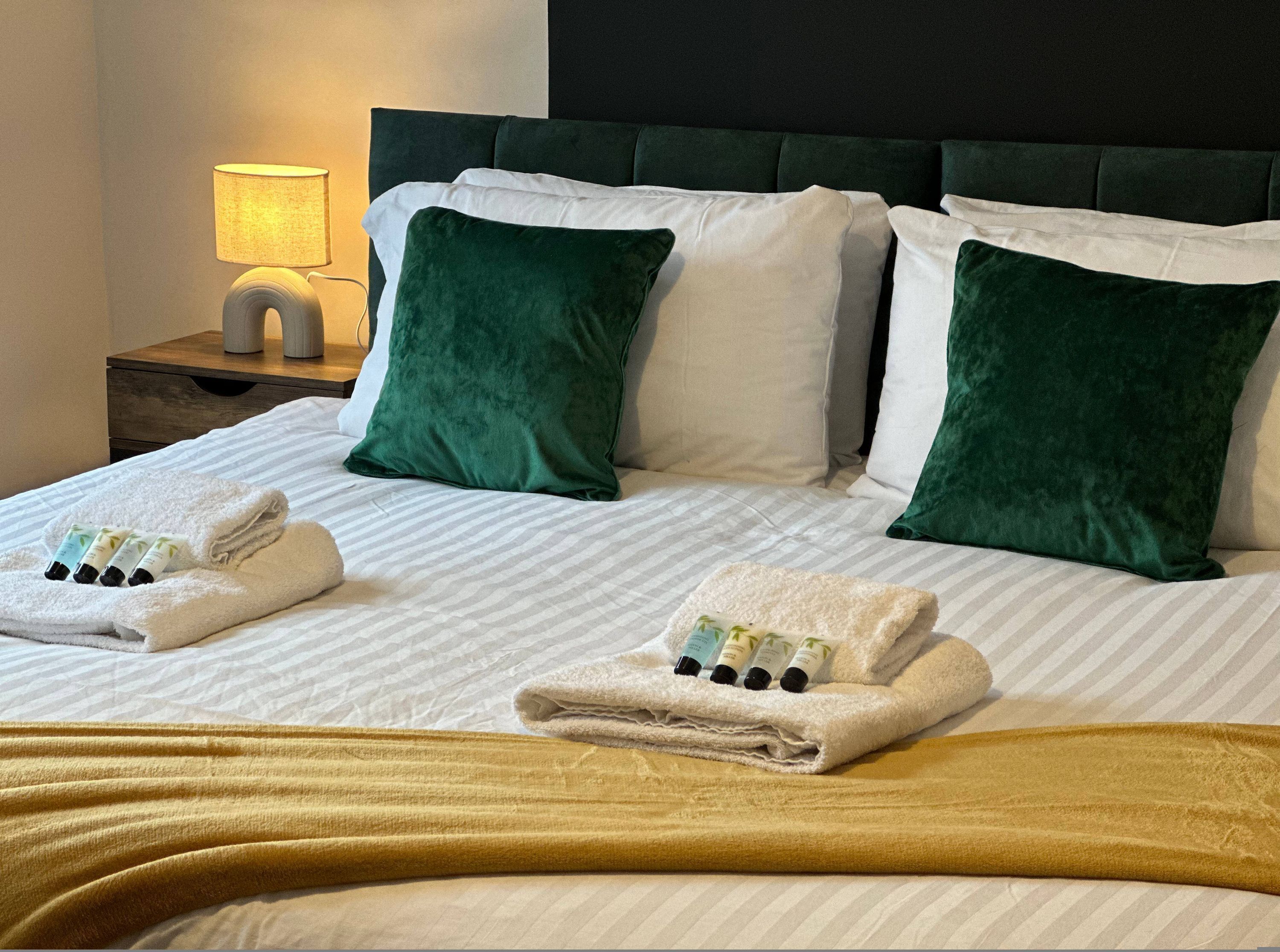 Una cama bien hecha con almohadas blancas y verdes, una toalla doblada y una manta amarilla.