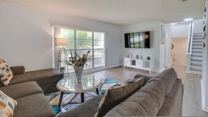 Una sala de estar moderna con un sofá, una mesa de centro con un florero, un televisor de pantalla plana montado en la pared y una escalera que sube a otro nivel.