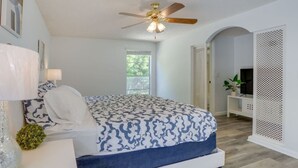Un dormitorio con cama, ventilador de techo, ventana y paredes blancas.