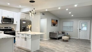 Une cuisine moderne avec des meubles blancs, un îlot central et un coin repas comprenant un canapé gris et un pouf.