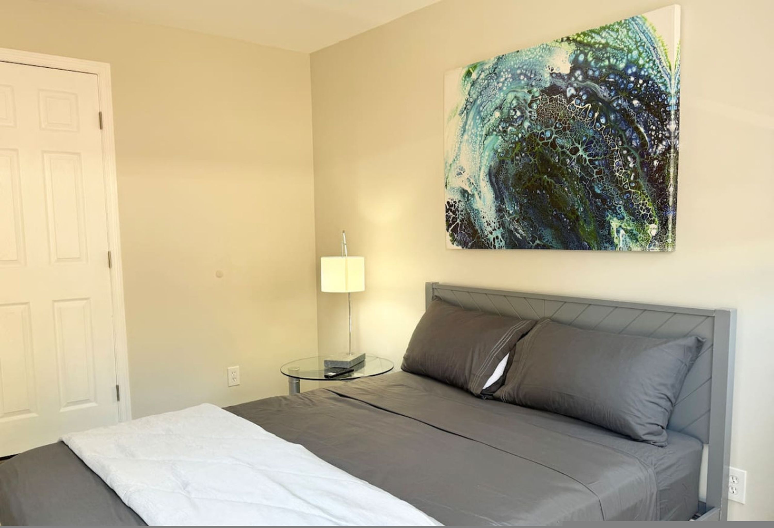Ein Schlafzimmer mit einem Bett, einem Nachttisch mit Lampe und einem an der Wand hängenden abstrakten Gemälde.