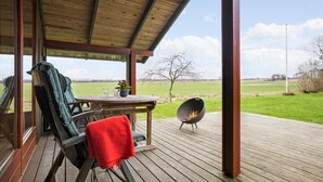 A covered outdoor area with a wooden deck, a round table, a chair, a grill, and a view of a grassy field. - 3 bedroom beautiful home in Hundested (Hundested)