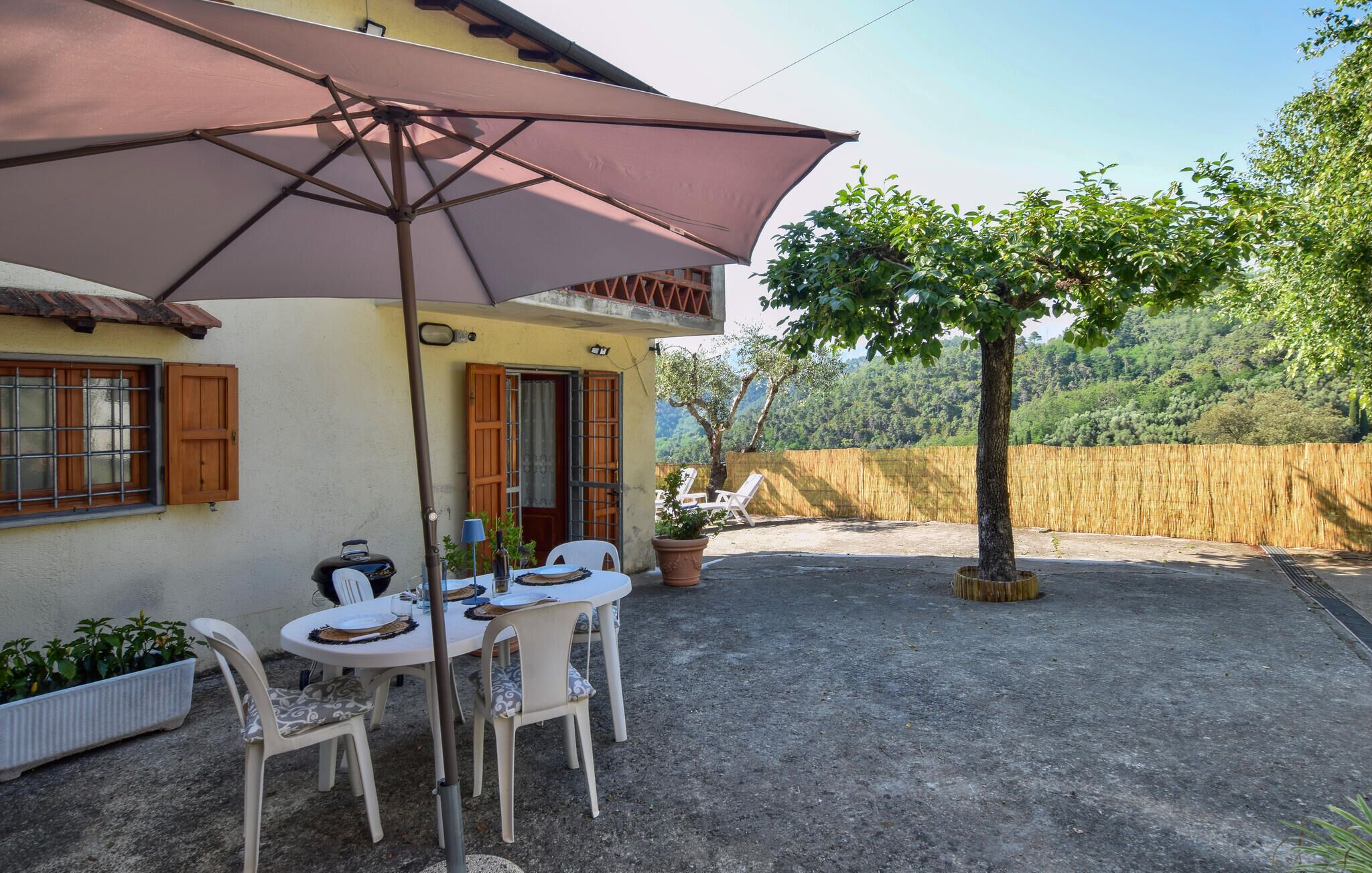 Um pátio com mesa e cadeiras, um grande guarda-sol e vista para uma encosta arborizada.
