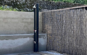 Una struttura nera verticale con un soffione doccia, un muro in cemento e un recinto in bambù.
