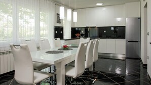 A modern kitchen with white cabinets, a stainless steel refrigerator, and a dining area with a white table and chairs. - Villa in Croatia Near Blue & Red Lakes (Imotski)