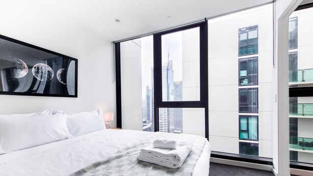 A modern hotel room with a large bed, a city view through floor-to-ceiling windows, and a framed artwork on the wall. - Relax & Recharge 1-Bedroom Apartment with Pool and Gym (Melbourne)