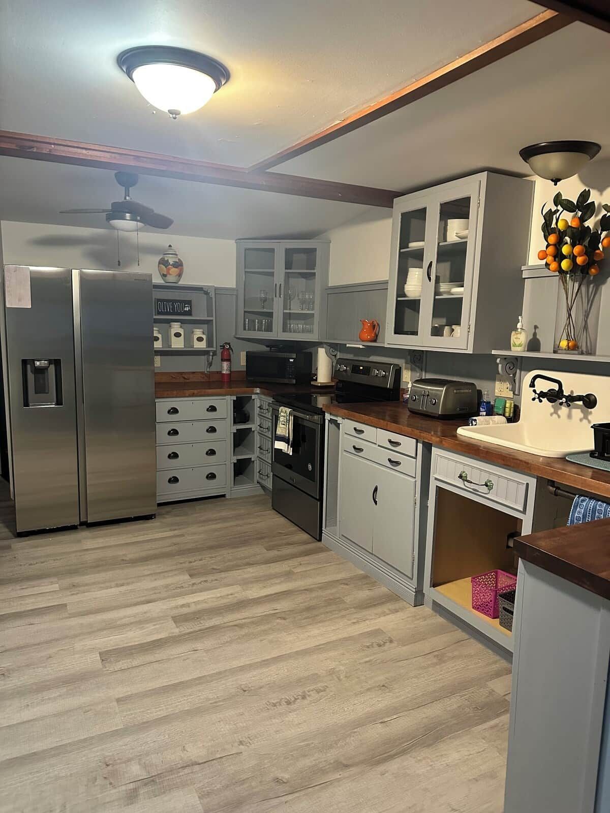 Una cocina moderna con un refrigerador de acero inoxidable, horno empotrado y microondas. Hay armarios con puertas de vidrio, una encimera de madera y una lámpara en el techo.
