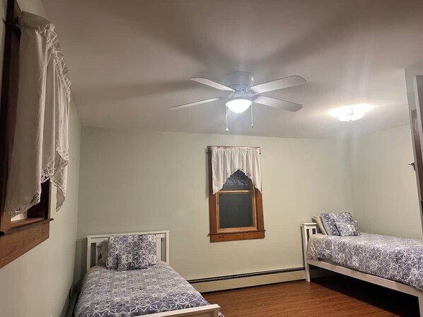 A bedroom with two single beds, a ceiling fan, a window with a curtain, and a wooden floor.
