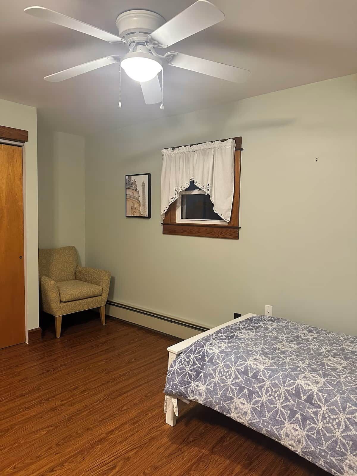 A bedroom with a bed, a chair, a ceiling fan, a window with a curtain, and a framed picture on the wall.