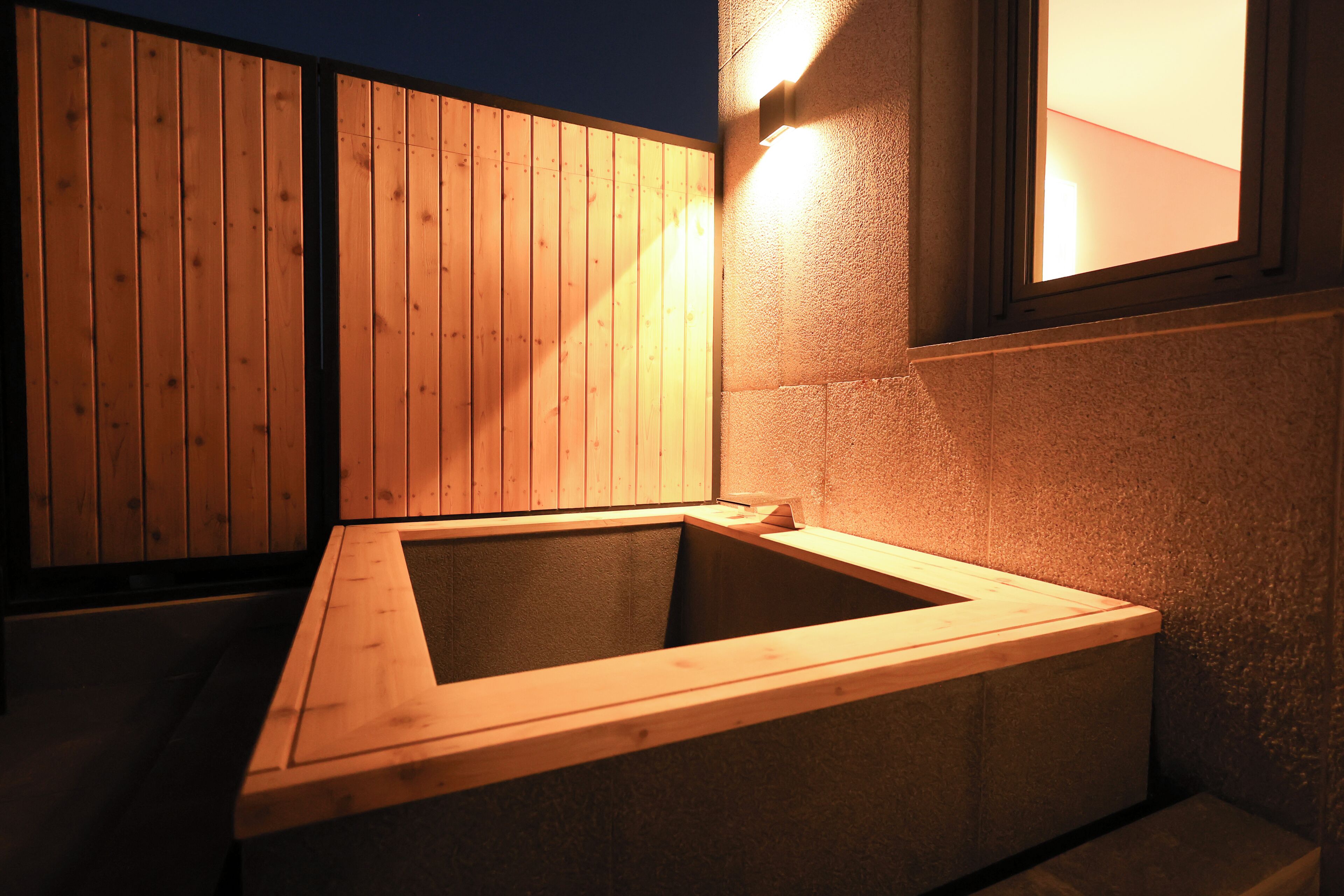 A wooden hot tub with a stone surround, a wooden fence, and a window with light.