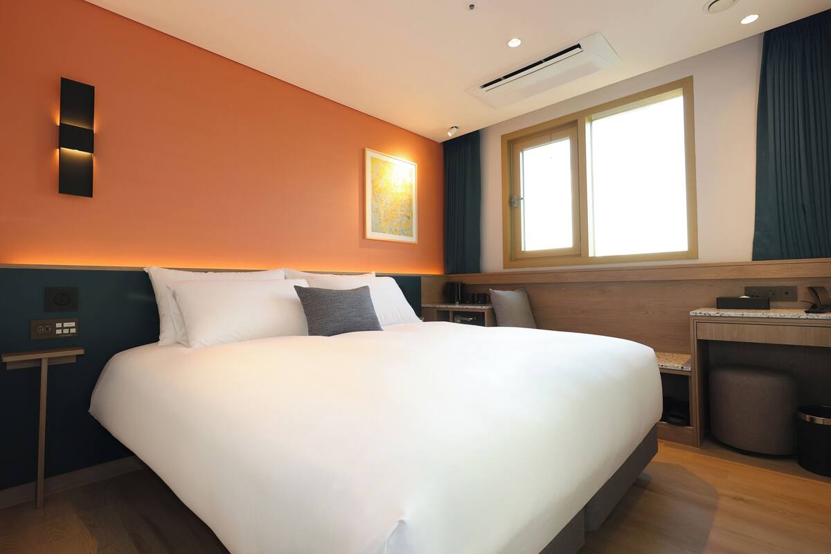 A hotel room with a large bed, a desk, a chair, a window with curtains, and an air conditioning unit.