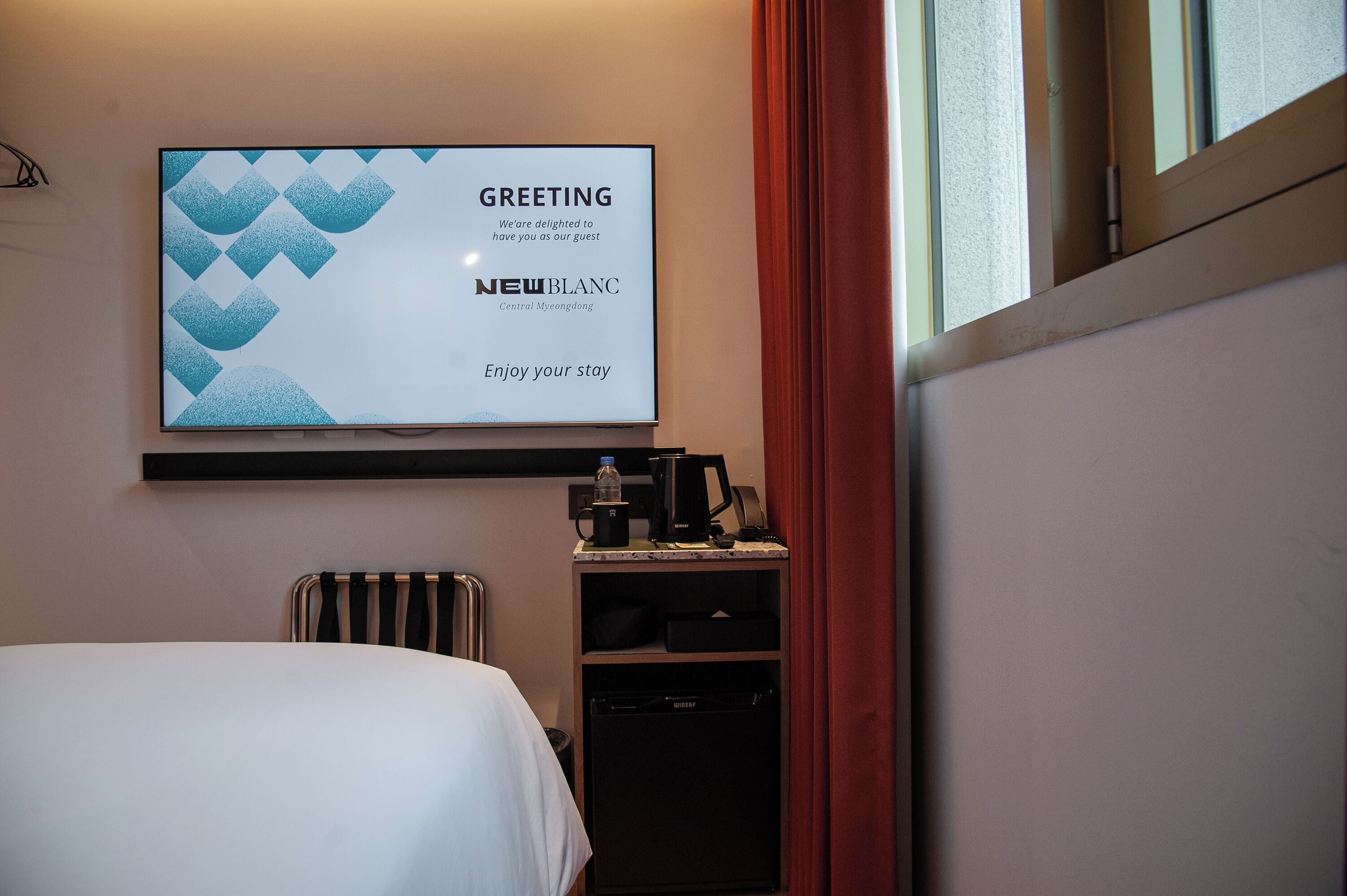 Una habitación de hotel con una cama, un televisor de pared mostrando un mensaje de bienvenida, una cafetera y un mueble auxiliar.