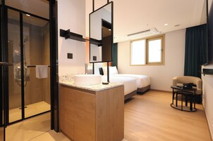 A modern hotel room with a glass-enclosed shower, a wooden vanity with a sink, a bed with white linens, and a small table with a chair.