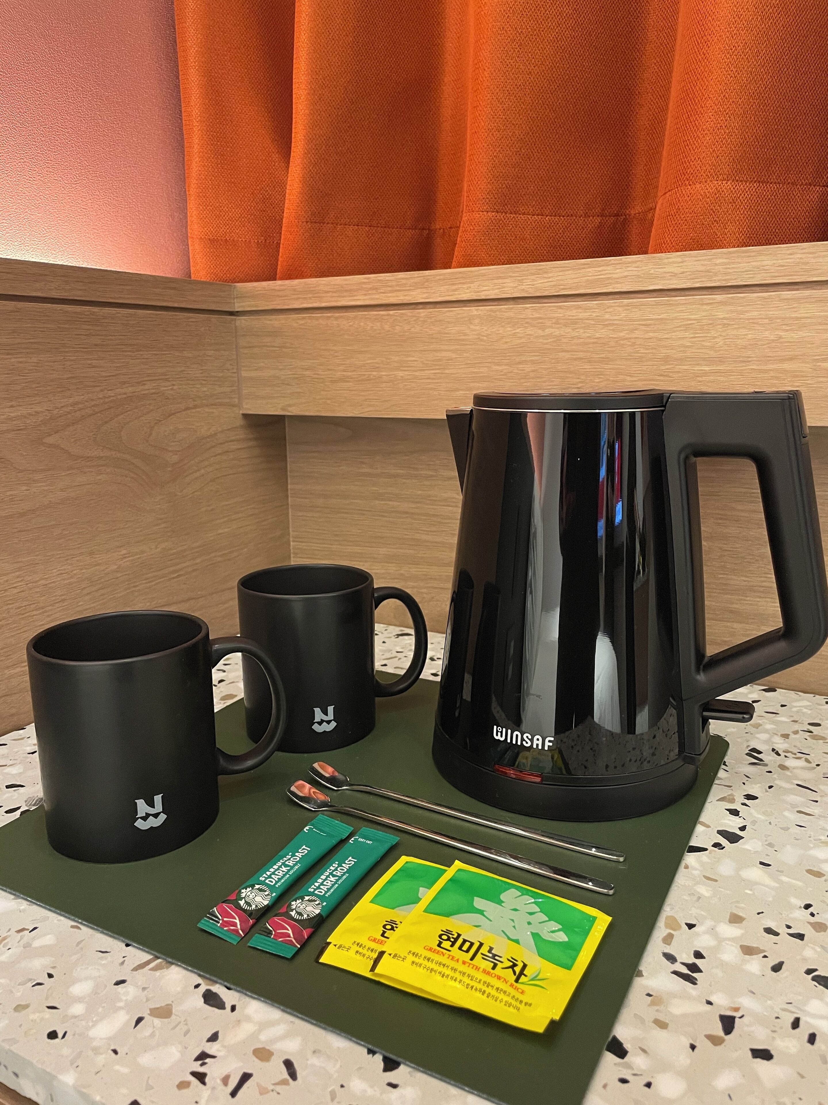 A kitchenette with a black coffee maker, two black mugs, a green tray, and two packets of tea.